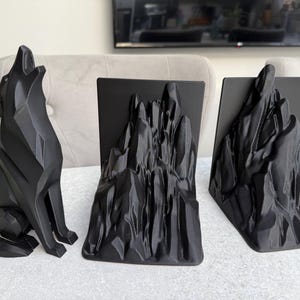 May include: A black geometric wolf statue howling, next to two black mountain-shaped bookends. The objects are on a white surface, creating a striking contrast. The bookends have a rectangular base.