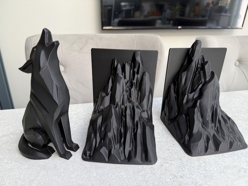 May include: A black geometric wolf statue howling, next to two black mountain-shaped bookends. The objects are on a white surface, creating a striking contrast. The bookends have a rectangular base.