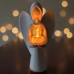 May include: A white ceramic angel figurine holding a glowing candle. The angel has outstretched wings and a serene expression. The candle emits a warm, yellow light, creating a soft glow. The background features blurred, out-of-focus lights.