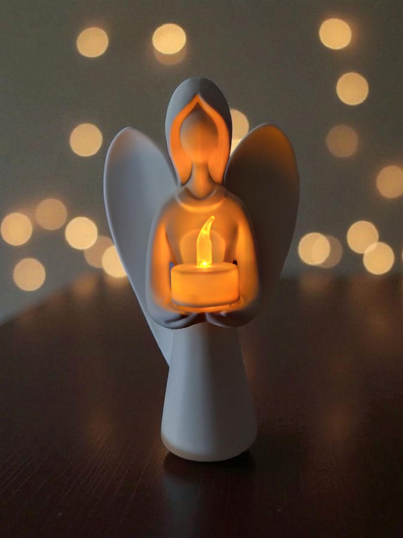 May include: A white ceramic angel figurine holding a glowing candle. The angel has outstretched wings and a serene expression. The candle emits a warm, yellow light, creating a soft glow. The background features blurred, out-of-focus lights.