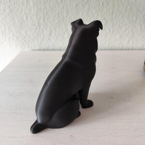 May include: A black dog figurine, viewed from the side and rear, sitting upright. The sculpture has a smooth, matte finish and is set against a neutral background. The dog's ears are perked up, and its tail is slightly raised.