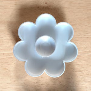 May include: A white, flower-shaped cookie cutter with a central circular design. The cutter is made of plastic and sits on a light brown wooden surface. The flower has six rounded petals.