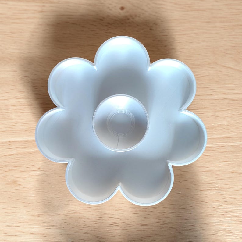 May include: A white, flower-shaped cookie cutter with a central circular design. The cutter is made of plastic and sits on a light brown wooden surface. The flower has six rounded petals.