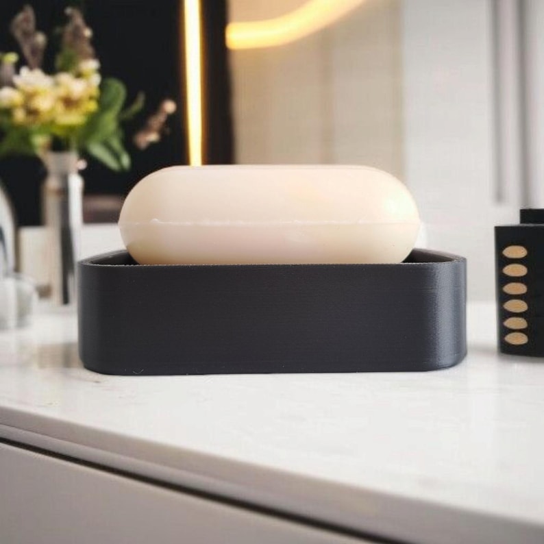 May include: A black soap dish with a bar of light beige soap. The soap dish is rectangular with rounded corners. The soap sits on a white countertop. A vase of flowers is in the background.