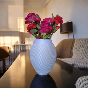 Puede incluir: Un jarr&oacute;n blanco texturizado con un dise&ntilde;o en espiral contiene un ramo de flores rosas y rojas. El jarr&oacute;n est&aacute; sobre una superficie oscura y reflectante. El fondo muestra un sal&oacute;n.