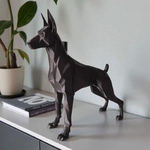 Op de afbeelding: Een donkergrijze geometrische hondensculptuur met puntige oren en een korte staart. De hond staat op een lichtgrijze ondergrond, met een plant en boeken op de achtergrond. De sculptuur heeft een modern, hoekig ontwerp.