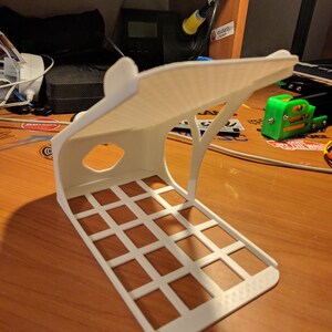 May include: A white 3D-printed phone stand with a grid base and an arched top. The stand has a rectangular shape with a cutout on the side. The stand is on a wooden surface.