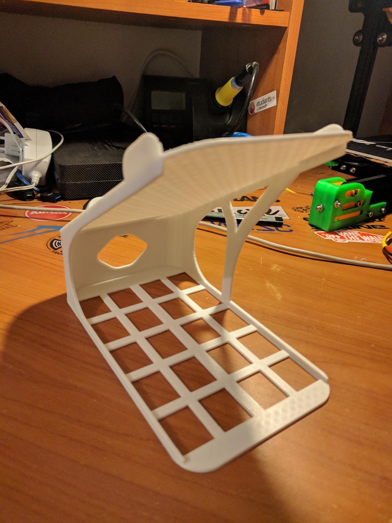May include: A white 3D-printed phone stand with a grid base and an arched top. The stand has a rectangular shape with a cutout on the side. The stand is on a wooden surface.