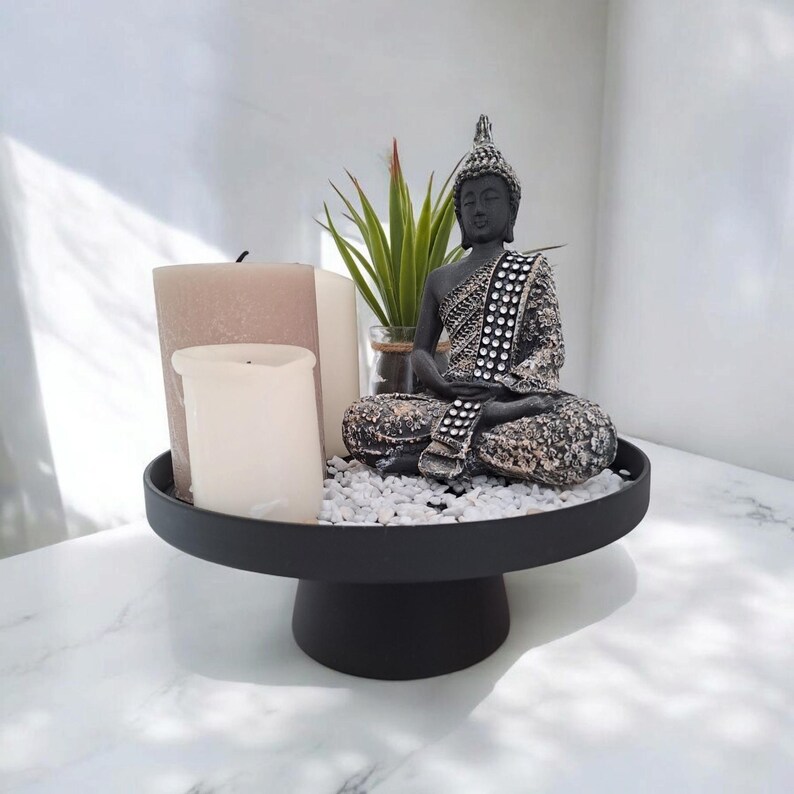 May include: A black decorative tray holds a seated Buddha statue, candles, and a small plant. The Buddha is grey with silver accents. The tray is on a white marble surface, creating a serene and zen-like atmosphere.