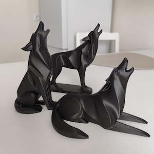 May include: Three black wolf sculptures in different poses, all with their heads raised and mouths open as if howling. One wolf is seated, one standing, and one lying down. The sculptures are on a white surface.