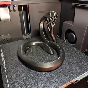 Puede incluir: Escultura en 3D de color gris oscuro que representa una serpiente enrollada con la cabeza levantada. La cabeza de la serpiente tiene un dise&ntilde;o detallado y texturizado. La escultura est&aacute; sobre una superficie negra texturizada.