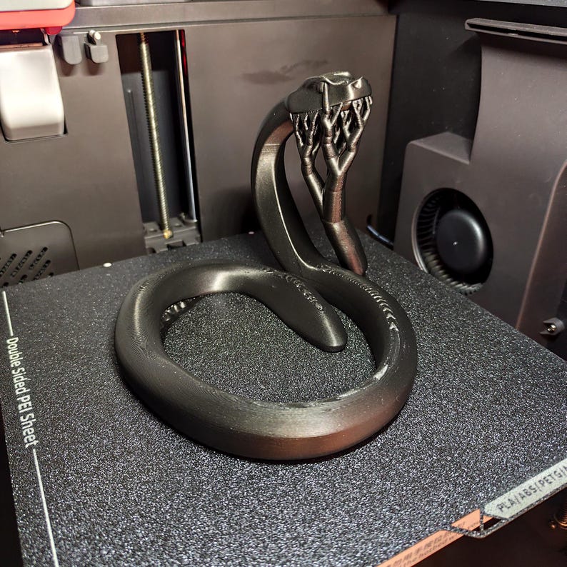 Puede incluir: Escultura en 3D de color gris oscuro que representa una serpiente enrollada con la cabeza levantada. La cabeza de la serpiente tiene un dise&ntilde;o detallado y texturizado. La escultura est&aacute; sobre una superficie negra texturizada.