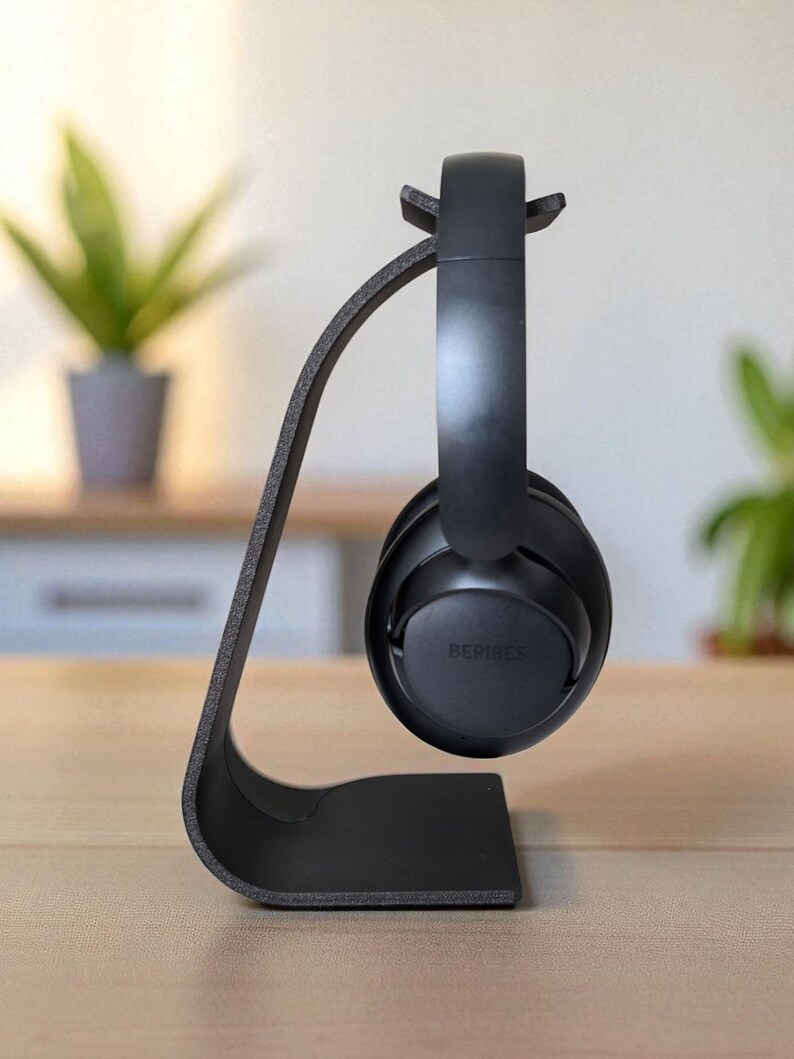 Peut inclure: Un support de casque noir au design incurv&eacute;, tenant une paire de casques noirs. Les &eacute;couteurs portent la marque "BERIBES". Le support est pos&eacute; sur une surface en bois, avec un arri&egrave;re-plan flou.