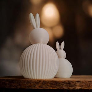 May include: Two white ceramic bunny figurines. The larger bunny has a ribbed body and a smooth head with upright ears. The smaller bunny has a smooth body and head with upright ears. Both are on a wooden surface.