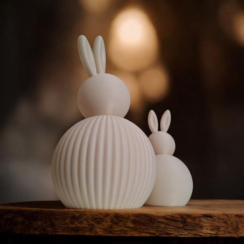 May include: Two white ceramic bunny figurines. The larger bunny has a ribbed body and a smooth head with upright ears. The smaller bunny has a smooth body and head with upright ears. Both are on a wooden surface.