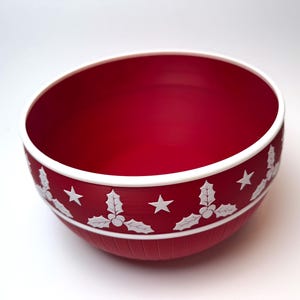 May include: A red bowl with a white rim and a decorative band featuring white holly and star designs. The bowl has a smooth interior and a textured exterior, perfect for holiday decor.