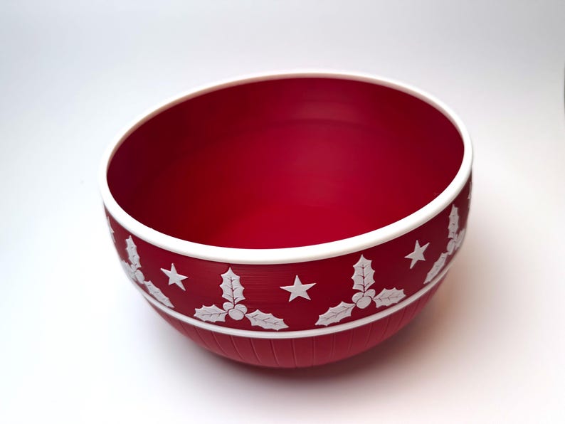 May include: A red bowl with a white rim and a decorative band featuring white holly and star designs. The bowl has a smooth interior and a textured exterior, perfect for holiday decor.
