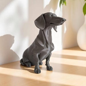 May include: A dark gray dachshund dog statue in a seated position. The sculpture has a smooth, matte finish and detailed features, including floppy ears and a long snout. The dog is positioned on a wooden surface, with a white wall in the background.
