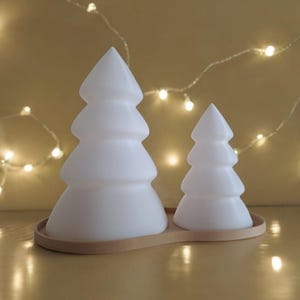 May include: Two white ceramic Christmas tree shaped candle holders on a beige base. The larger tree is in the background and the smaller tree is in the foreground.