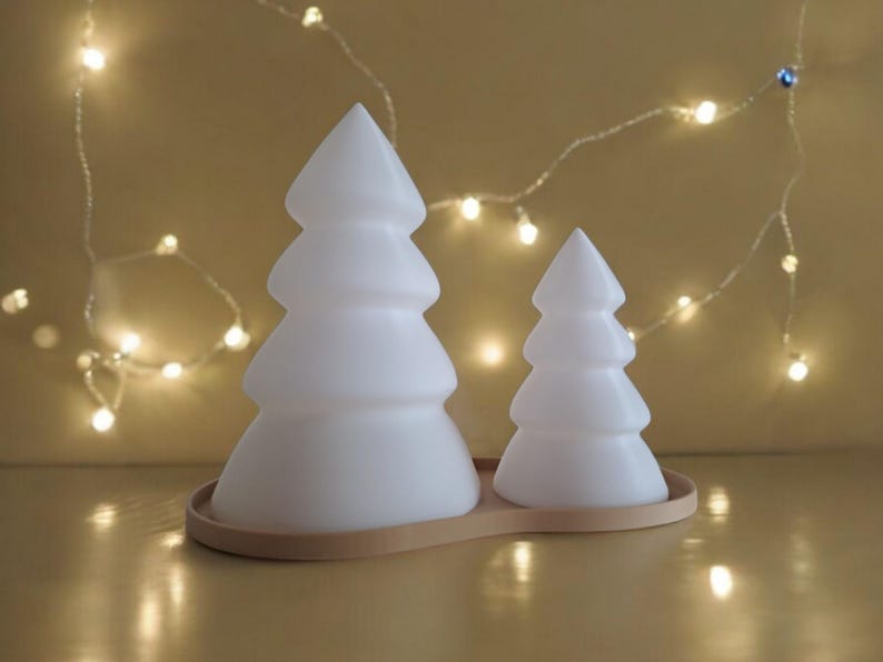 May include: Two white ceramic Christmas tree shaped candle holders on a beige base. The larger tree is in the background and the smaller tree is in the foreground.