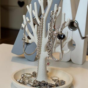 May include: A white, tree-shaped jewelry organizer with various silver necklaces, bracelets, and rings. The organizer has a round base and multiple branches to hang jewelry. A gray decorative object is behind the organizer.