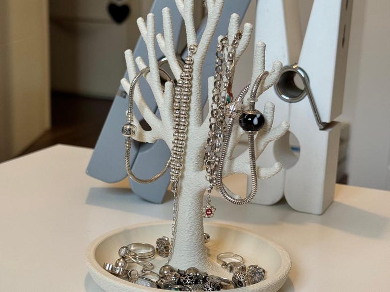 May include: A white, tree-shaped jewelry organizer with various silver necklaces, bracelets, and rings. The organizer has a round base and multiple branches to hang jewelry. A gray decorative object is behind the organizer.