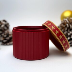 May include: A red, cylindrical container with a lid decorated with gold stars. The container has a ribbed texture. The lid is slightly ajar, revealing the interior. The background includes pine cones and a gold bauble, suggesting a festive theme.
