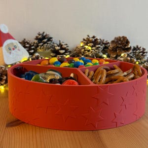 May include: A red, round serving tray with star-shaped cutouts, filled with colorful candies and pretzels. A Santa Claus decoration and pine cones are in the background, suggesting a holiday theme.