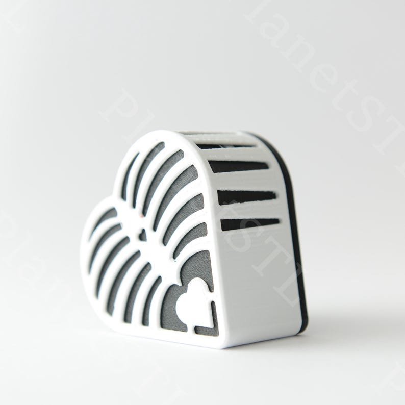 May include: A white, heart-shaped decorative object with a black interior. The object has a slotted design on the front and top, with a small heart-shaped cutout on the side. The object is set against a white background.