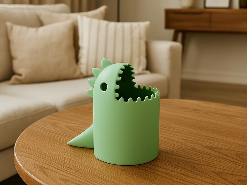 May include: A light green dinosaur-shaped container with a wide open mouth and a small tail. The container is cylindrical with a jagged edge around the opening. The dinosaur has a small hole on the side.