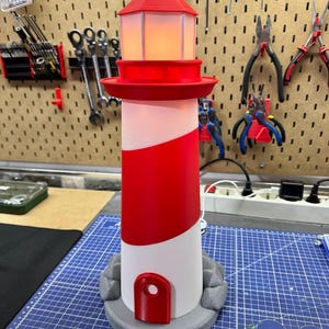 May include: A red and white lighthouse lamp with a light inside. The lamp has a red top and a red stripe around the middle. The base is gray and has a door. The lamp is approximately 12 inches tall.