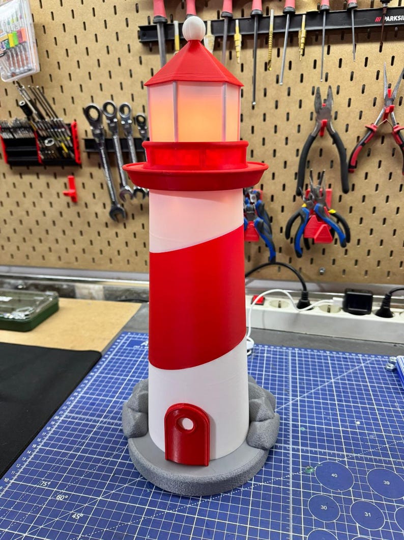 May include: A red and white lighthouse lamp with a light inside. The lamp has a red top and a red stripe around the middle. The base is gray and has a door. The lamp is approximately 12 inches tall.