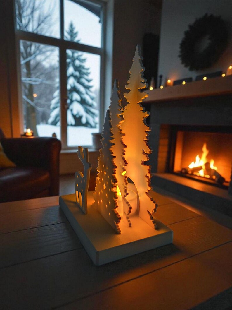 May include: A white, laser-cut Christmas tree and reindeer candle holder on a wooden table. The candle holder has a warm glow from the lit candle inside. A cozy fireplace and snowy window are in the background, creating a festive holiday scene.