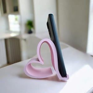 May include: A pink heart-shaped phone stand holds a black smartphone in a vertical position. The stand is on a white surface.
