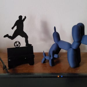 May include: A black silhouette of a soccer player kicking a ball, and two blue balloon dog sculptures. The soccer player is on a black stand. The balloon dogs are in different sizes and poses, all on a wooden surface.