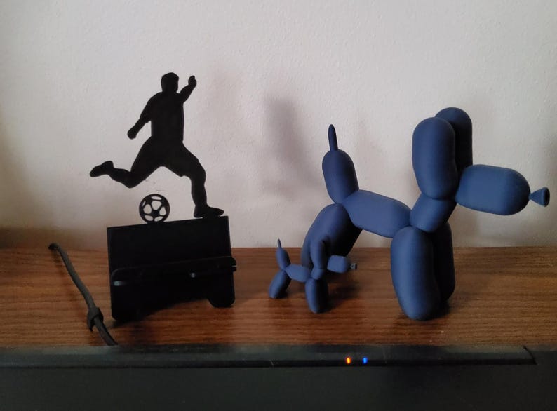 May include: A black silhouette of a soccer player kicking a ball, and two blue balloon dog sculptures. The soccer player is on a black stand. The balloon dogs are in different sizes and poses, all on a wooden surface.