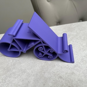 May include: A purple phone stand with a unique design. The stand has a curved base with decorative swirls and a slanted back for device support. The word "SK" is visible on the stand. The stand is made of plastic.