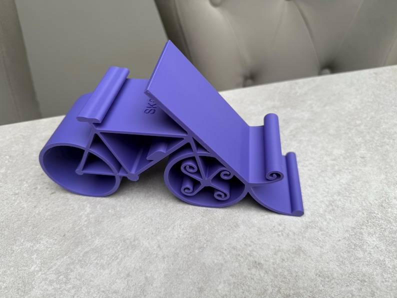 May include: A purple phone stand with a unique design. The stand has a curved base with decorative swirls and a slanted back for device support. The word "SK" is visible on the stand. The stand is made of plastic.