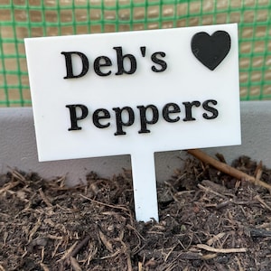 Puede incluir: Un marcador de plantas blanco con el texto "Deb's Peppers" en negro, coronado por un coraz&oacute;n negro. El marcador est&aacute; insertado en tierra oscura, lo que sugiere un jard&iacute;n. Se ve una malla verde en el fondo.