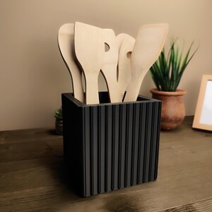 May include: A black, ridged utensil holder filled with wooden kitchen utensils. The utensils include spatulas and spoons. A small potted plant is visible in the background.
