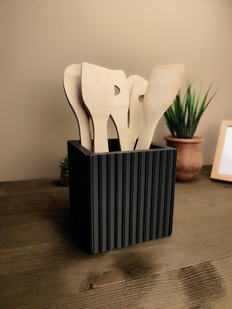 May include: A black, ridged utensil holder filled with wooden kitchen utensils. The utensils include spatulas and spoons. A small potted plant is visible in the background.