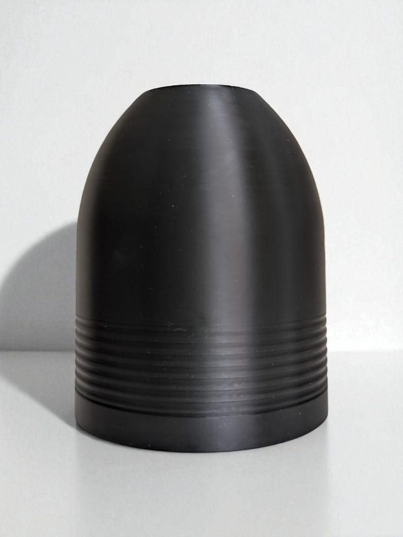 May include: A matte black, bullet-shaped object with a rounded top and a series of horizontal, grooved bands near the base. The object is set against a plain white background, with a shadow to the left.