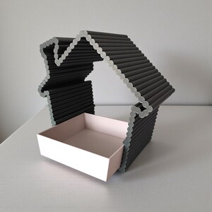 May include: A decorative house-shaped shelf with a black exterior and a light pink interior drawer. The house frame is constructed from numerous parallel black rods, with a silver-colored outline. The drawer is rectangular and sits at the base of the house.