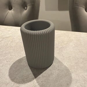 May include: A gray, cylindrical vase with vertical ribbed detailing. The vase is made of a solid material and sits on a light-colored surface. The vase casts a shadow on the surface.