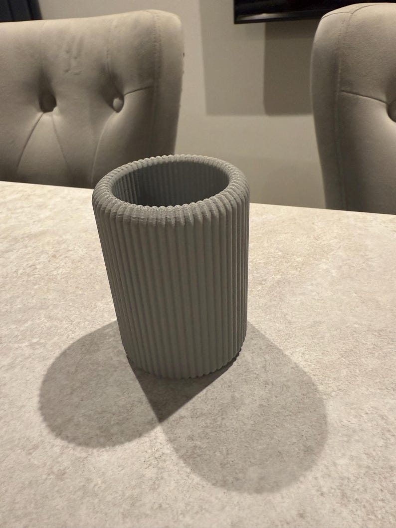 May include: A gray, cylindrical vase with vertical ribbed detailing. The vase is made of a solid material and sits on a light-colored surface. The vase casts a shadow on the surface.