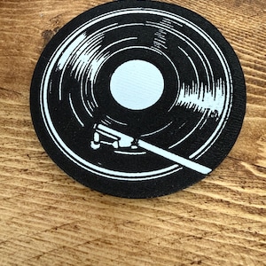 May include: A black and white circular coaster featuring a vinyl record design. The coaster has a white center and a white tonearm graphic. The design is printed on a black background.