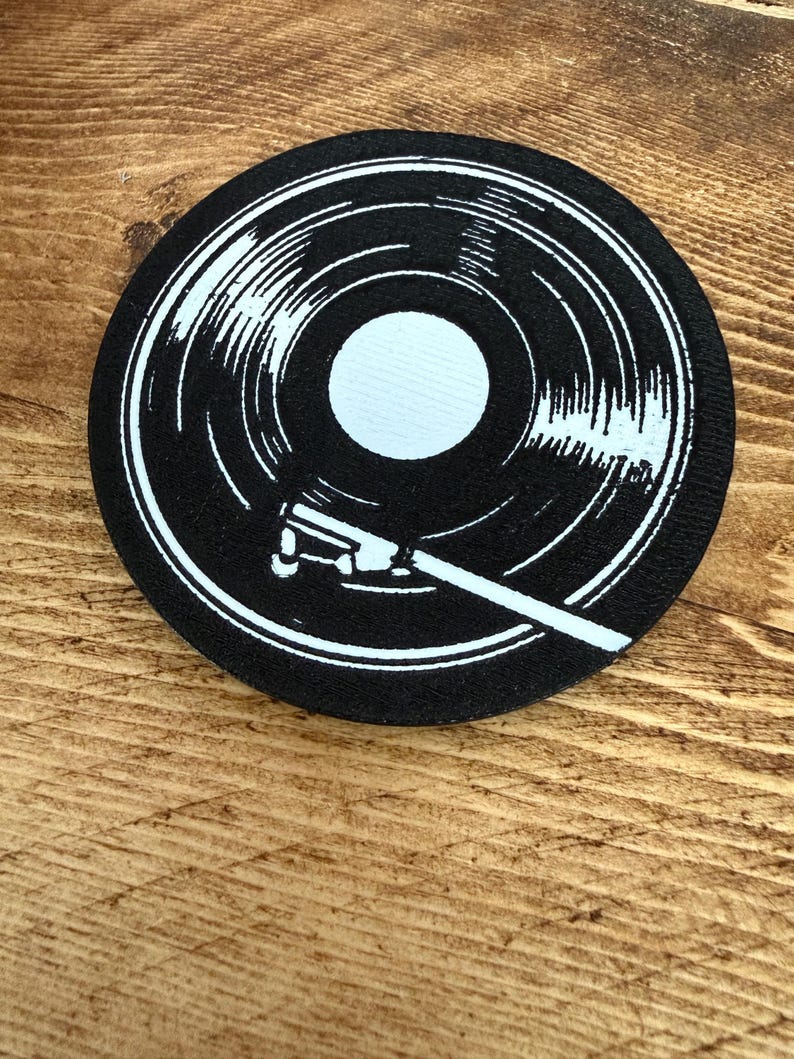 May include: A black and white circular coaster featuring a vinyl record design. The coaster has a white center and a white tonearm graphic. The design is printed on a black background.