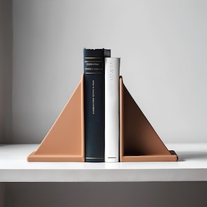 Op de afbeelding: Een paar terracotta-kleurige driehoekige boekensteunen houden twee boeken vast. E&eacute;n boek is marineblauw met gouden letters, de andere is wit. De boekensteunen staan op een witte plank tegen een neutrale achtergrond.
