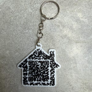 May include: White house-shaped keychain with a black QR code design. The keychain has a silver chain and ring for attaching to keys or bags. The house shape includes a chimney.