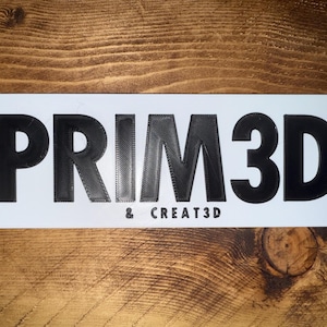 Pu&ograve; includere: Un cartello rettangolare bianco con la scritta "PRIM3D" in grandi lettere nere stampate in 3D. Sotto, il testo pi&ugrave; piccolo recita "& CREATE3D". Il cartello &egrave; posto su uno sfondo di legno.